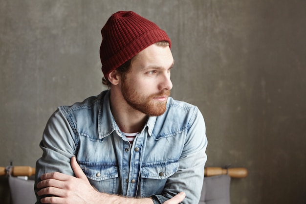 Indoor shot of handsome bearded Caucasian hipster wearing maroon hat and denim shirt sitting at cafe, looking away with thoughtful expression on his face, wondering what future has in store for him