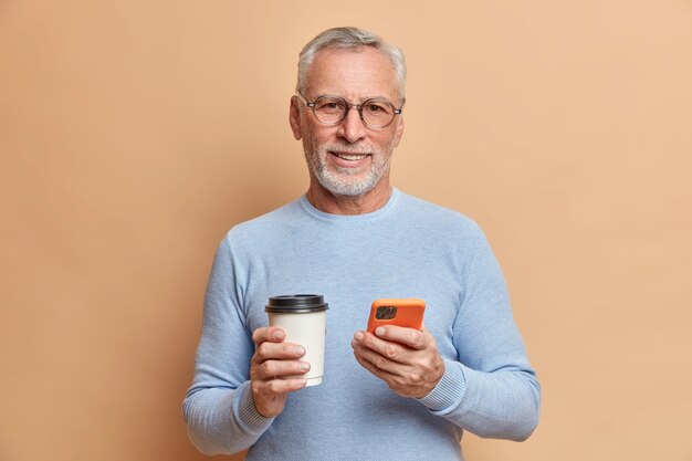 Indoor shot of handsome bearded mature man has free time after work checks social networks on smartphone drinks takeaway coffee wears spectacles and blue jumper isolated over brown wall
