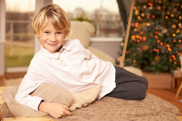 Крытый снимок красивого симпатичного десятилетнего мальчика с аккуратной стрижкой и радостной улыбкой, позирующего на подушке, лежащего на полу перед елкой, украшенной игрушками и гирляндой. Детство и праздники