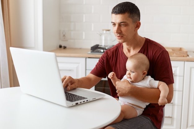 Indoor shot of handsome man with dark hair wearing burgundy casual t shirt, working on laptop while babysitting, looking at notebook screen posing in white kitchen.