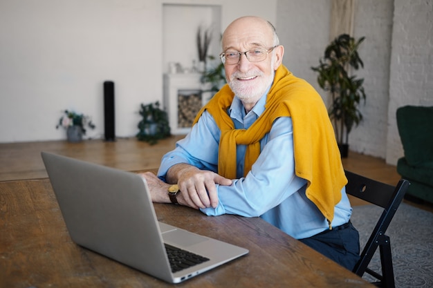 Снимок в помещении красивого позитивного небритого шестидесятилетнего писателя в очках и стильной одежде, который работает удаленно, сидит за столом перед открытым портативным компьютером и широко улыбается