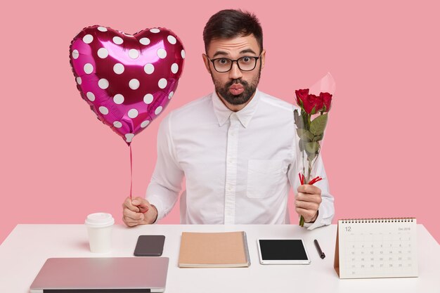 Indoor shot of handsome unshaven man pouts lips, carries valentine and bouquet, has romantic relationships in office, poses over pink studio wall