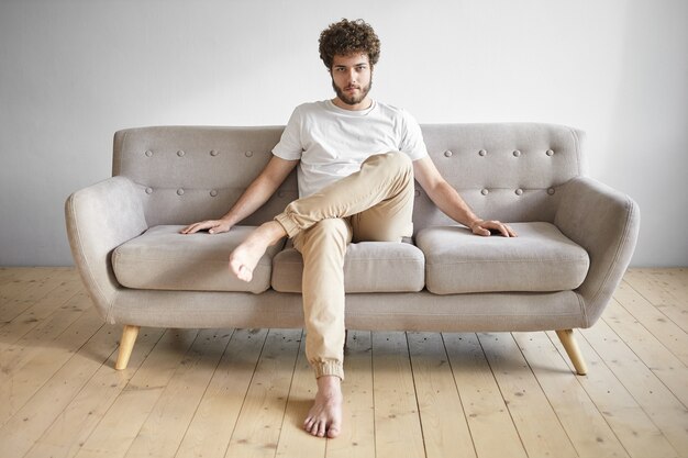 Indoor shot of handsome young bearded man wearing white t-shirt and blue jeans sitting barefooted on comfortable gray couch,  and smiling, blank copyspace wall