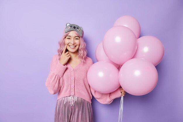 Indoor shot of happy Asian woman enjoys festive occasion smiles positively wears blindfold jumper and pleated skirt holds bunch of inflated balloons poses against purple background Holidays concept