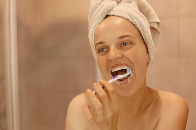 Indoor shot of happy attractive female brushing her teeth, having hygiene and beauty procedures at home in bathroom, standing with white towel on her head.