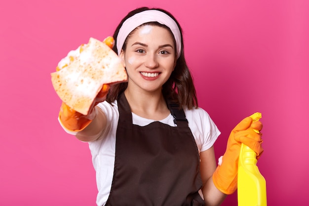 Внутренний снимок счастливой кавказской молодой женщины в белой повседневной футболке, ободке для волос, коричневом фартуке, держит губку и моющее средство, готов сделать домашнюю работу, стоит улыбается на розовой стене. Концепция гигиены