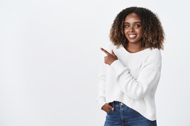 Indoor shot of happy charming beautiful dark-skinned female in sweater with curly hair pointing at upper left corner and smiling broadly