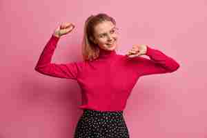 Free photo indoor shot of happy cheerful girl with pony tail dances to favorite music, raises hands, feels life energy and freedom, wears spectacles, red turtleneck, receives pleasure