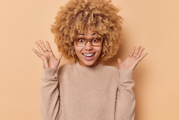 Indoor shot of happy curly haired woman feels excited keeps palms raised reacts on something amazing wears spectacles and casual jumper isolated over beige background Human reactions concept