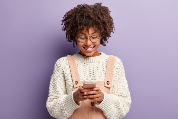 Tiro al coperto di donna riccia felice con l'espressione facciale soddisfatta, legge un bel messaggio dal ragazzo, concentrato nello smartphone