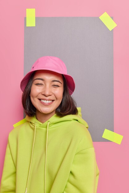 幸せなかわいいアジアの10代の少女の笑顔の屋内ショットは、ピンクの壁の漆喰紙に対してカジュアルな服のポーズを着て正面で面白い会話のにやにや笑いを楽しんでいます