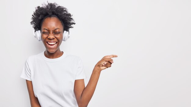 Indoor shot of happy dark skinned woman laughs happily indicates away on blank space