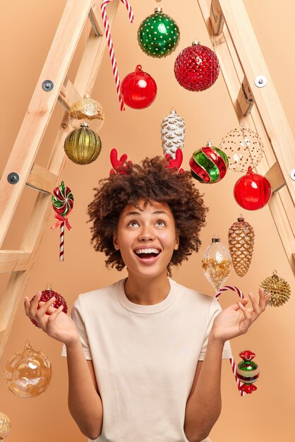 Indoor shot of happy dark skinned woman looks gladfully above spreads palms and smiles broadly wears red reindeer horns uses ladder to decorate house for new year surrounded by christmas toys