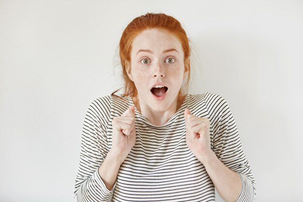 Indoor shot of happy ecstatic young woman with freckles having shocked look, exclaiming, keeping mouth wide open and fists clenched while celebrating success, surprised with her unexpected victory