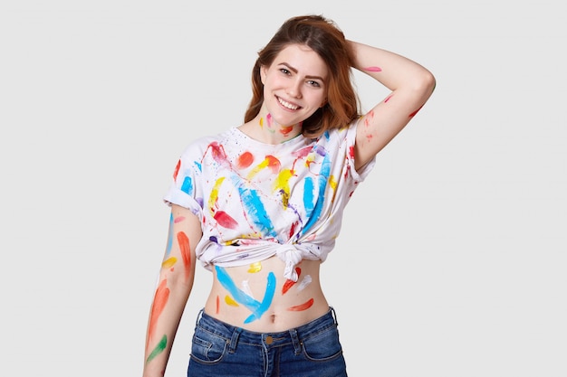 Indoor shot of happy european female painter has dirty body and white t shirt with colourful paints, shows belly, keeps hand behind head, isolated over white wall creats artwork or masterpiece