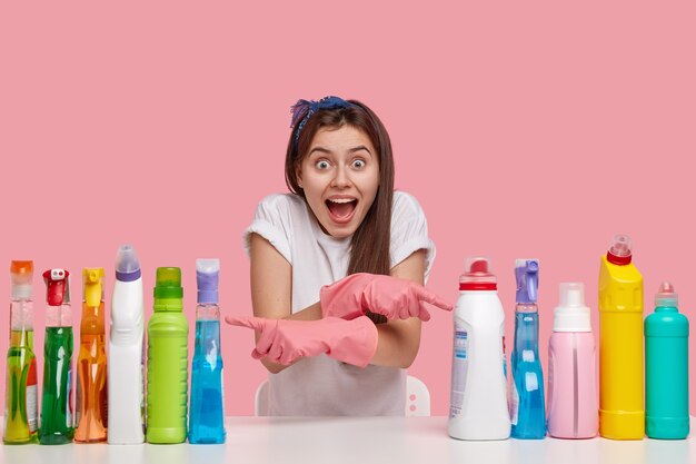 Indoor shot of happy European lady with overjoyed expression, keeps mouth opened, crosses hand and indicates at two sides on detergents