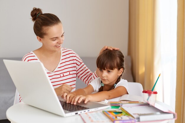 Indoor shot of happy family doing homework together, sitting at table in living room, mommy praises her daughter for school success, caress her little daughter, touching her head.