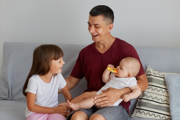 Indoor shot of happy family, smiling father playing with two daughters on sofa, cute baby biting toy, dad spending time with charming children at home.
