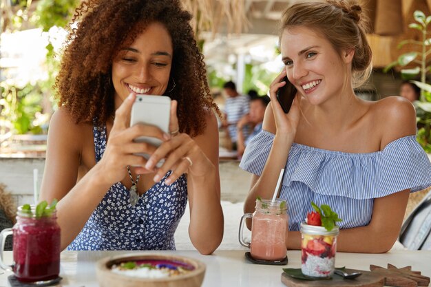 幸せな女性の屋内ショットは、現代のスマートフォンを使用し、ソーシャルネットワークをサーフィンし、モバイルで会話し、カフェテリアで自由な時間を過ごし、スムージーを飲みます。うれしそうな女性は夏休み中に再現します