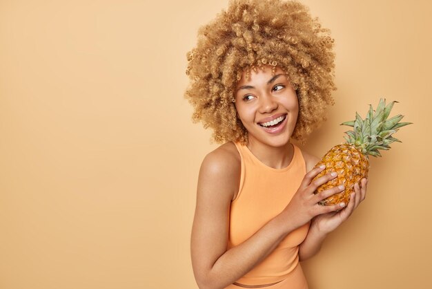 Снимок в помещении счастливой милой женщины с вьющимися волосами держит любимые экзотические фрукты, такие как ананасы, с веселым выражением лица, смотрит в сторону, одетая в футболку, изолированную на коричневом фоне пустого пространства
