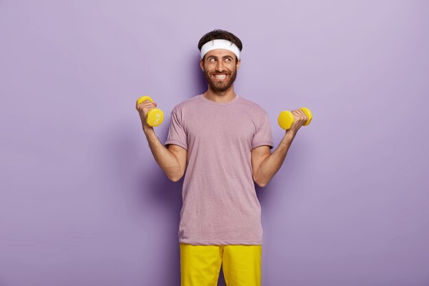 Indoor shot of happy man with bristle, raises two arms with weights, dressed in casual outfit, trains on biceps