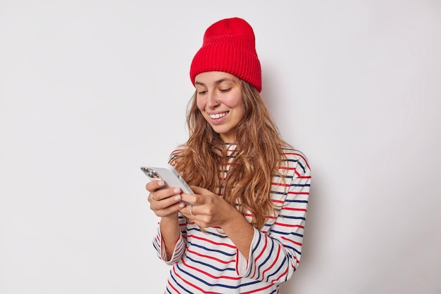 Снимок в помещении счастливой миллениальной девушки, отправляющей текстовые сообщения через приложение для смартфонов, с пирсингом в носу, одетой в повседневную одежду, изолированную на белом. Концепция современных технологий