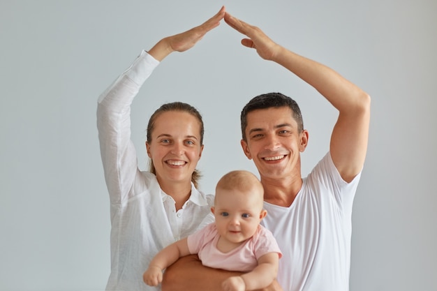 Indoor shot of happy positive family posing isolated light background, looking at camera, holding baby in hands, mom and dad making roof figure with hands arms over heads, safety.