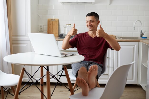 행복한 긍정적인 남자가 부엌 테이블에 앉아 웃고 있는 랩탑 디스플레이를 보고 엄지손가락을 치켜들고 새로운 프로젝트에 대한 고용주의 아이디어를 승인하는 실내 사진.