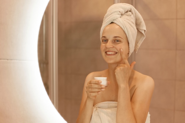 Colpo al coperto di una donna sorridente felice in piedi davanti allo specchio che si strofina la crema cosmetica sul viso, mettendo una crema idratante sulla pelle del viso in bagno.