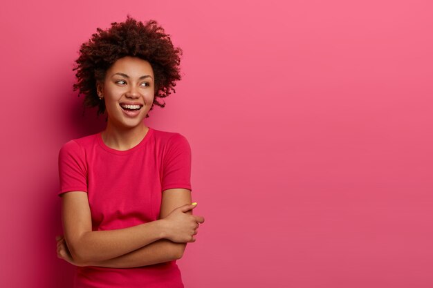 Indoor shot of happy woman keeps arms crossed over body, concentrated aside with broad smile, enjoys casual talk with someone, discusses something funny, poses in rosy . Happiness concept