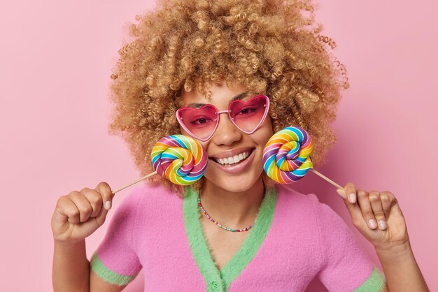 Indoor shot of happy woman with curly hair holds two colorful candies near face smiles toothily wears pink sunglasses and casual t shirt isolated over pink background Sweet tooth and fun concept