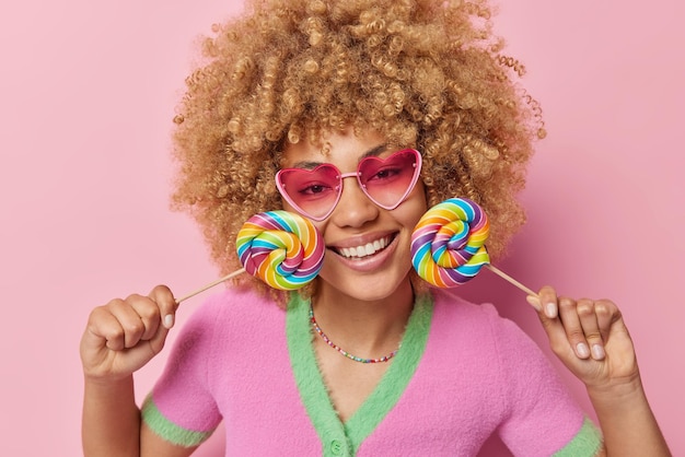 Снимок счастливой женщины с вьющимися волосами в помещении держит две разноцветные конфеты возле лица, зубасто улыбается, носит розовые солнцезащитные очки и повседневную футболку, изолированную на розовом фоне Сладкоежка и веселая концепция