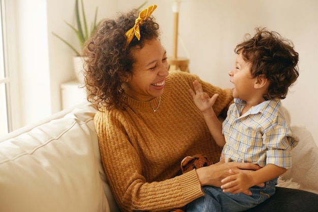 彼女の愛らしい幼児の息子を抱きしめて家でリラックスしている茶色のウェーブのかかった髪を持つ幸せな若いヒスパニック系女性の屋内ショット。陽気な母親が幼児の息子と絆を結ぶ、リビングルームのソファに座って、笑う