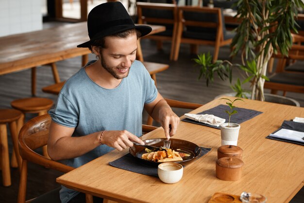 Indoor shot of happy young student wearing stylish hat eating delicious food with knife and fork during break at university canteen, enjoying fresh and healthy lunch