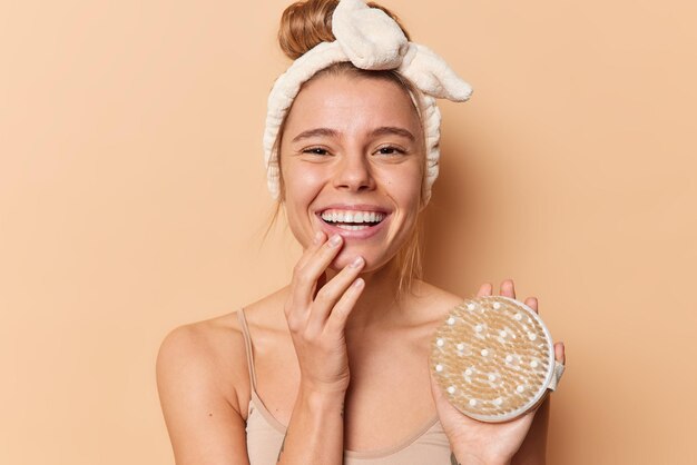 Indoor shot of happy young woman with clean glowing skin holds bath brush feels good has bare shoulders white teeth isolated over brown background. People hygiene and daily routines concept.