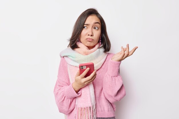 Indoor shot of hesitant young Asian woman shrugs shoulders with confused expression uses mobile phone for chatting onile wears pink jumper and scarf around neck isolated over white background