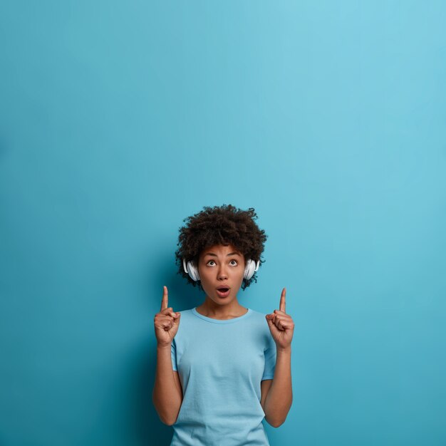 Indoor shot of impressed dark skinned woman wears headphones on ears, looks suprisingly above, indicates at blank space, shocked with stunning advertisement, isolated on blue wall. Music concept