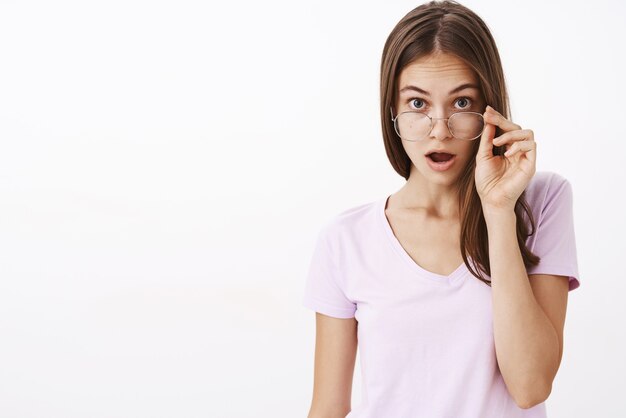 Indoor shot of impressed surprised attractive adult female brunette taking off glasses and dropping jaw raising eyebrows from wonder and amazement finding out new details of gossip over white wall