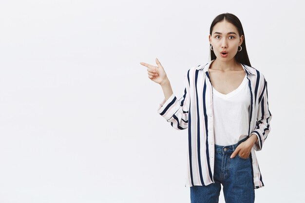 Indoor shot of impressed and surprised charming female student in stylish striped blouse, holding hand in pocket and pointing left with forefinger, gasping from amazement, standing over gray wall