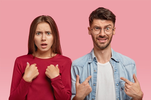Free photo indoor shot of indignant caucasian young woman and man point at themselves