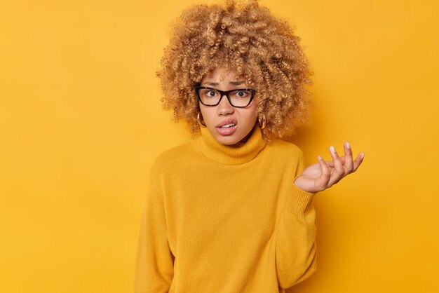 Indoor shot of indignant curly haired woman shrugs shoulders raises palm with displeased expression purses lips wears spectacles and jumper isolated over vivid yellow background feels indecisive.