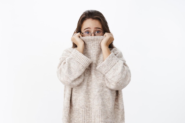 Free photo indoor shot of insecure worried young cute woman in glasses with brown hair pulling collar of sweater on nose and frowning nervously