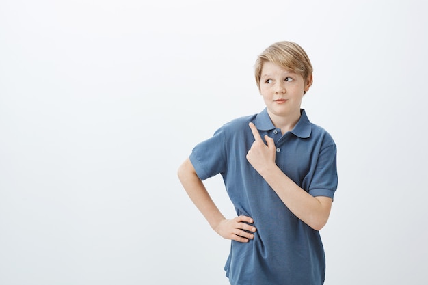Indoor shot of intrigued cute young boy with blond hair, lifting eyebrows with curious expression, holding hand on hip