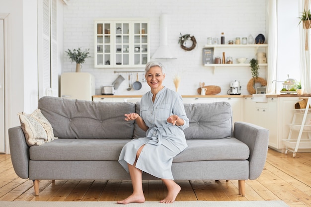 Indoor shot of joyful charming elderly housewife in stylish dress sitting on large gray couch with bare feet on floor with beaming smile, gesturing emotionally, being in good mood