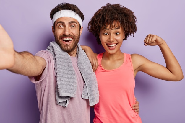 Indoor shot of joyful diverse couple keep muscle flexible, have daily workout, wear sports clothing stand closely look at camera with happy expression