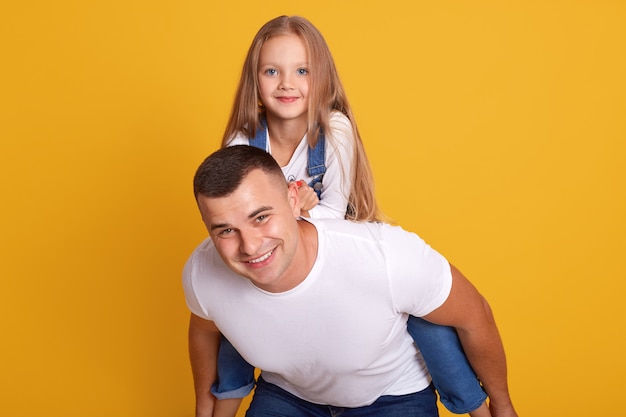 Indoor shot of joyful father giving piggyback ride to his daughter against yellow, happy family wearing casual clothing