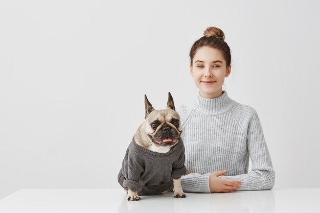 Indoor shot of joyful female pet lover with brown hair in bun. Housewife posing with her pedigree dog dressed in jumper sitting at table taking pleasure together. Positive human expressions