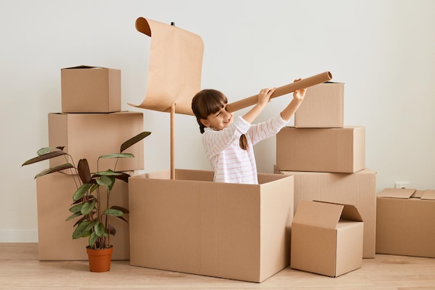 Indoor shot of little girl with dark hair and braids sitting in handmade ship from cardboard boxes and looking in paper spyglass child playing during relocating