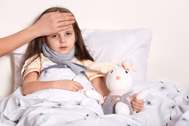 Indoor shot of little girl with fair hair lying in her bed, hugging favourite toy, having unknown hand on forehead, checking temperature