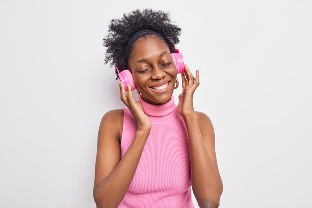 Indoor shot of lovely dark skinned woman closes eyes from pleasure listens to  favorite music keeps hands on stereo pink headphones 